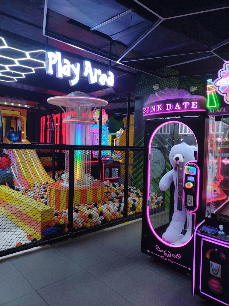 Neon-lit arcade claw machine with white bear prize next to ball pit and colorful slide at Sector 12 Faridabad play zone