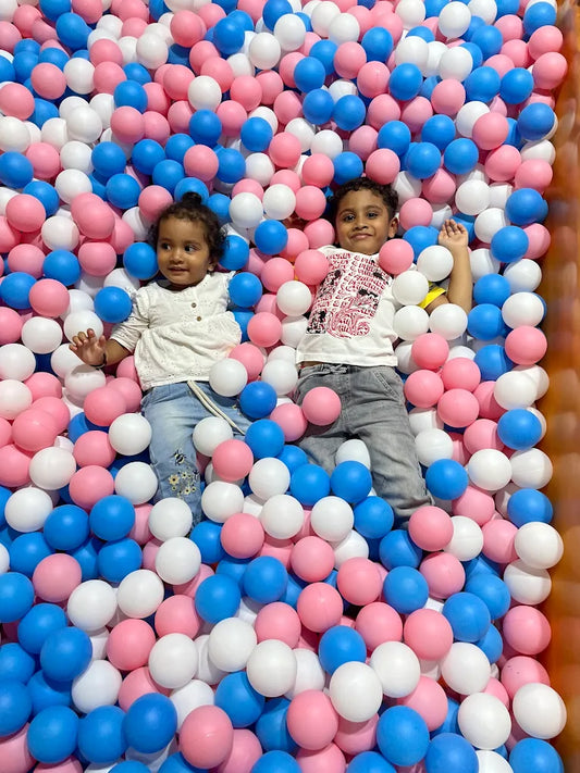 Two young children lie in a ball pit filled with pink, blue, and white balls.