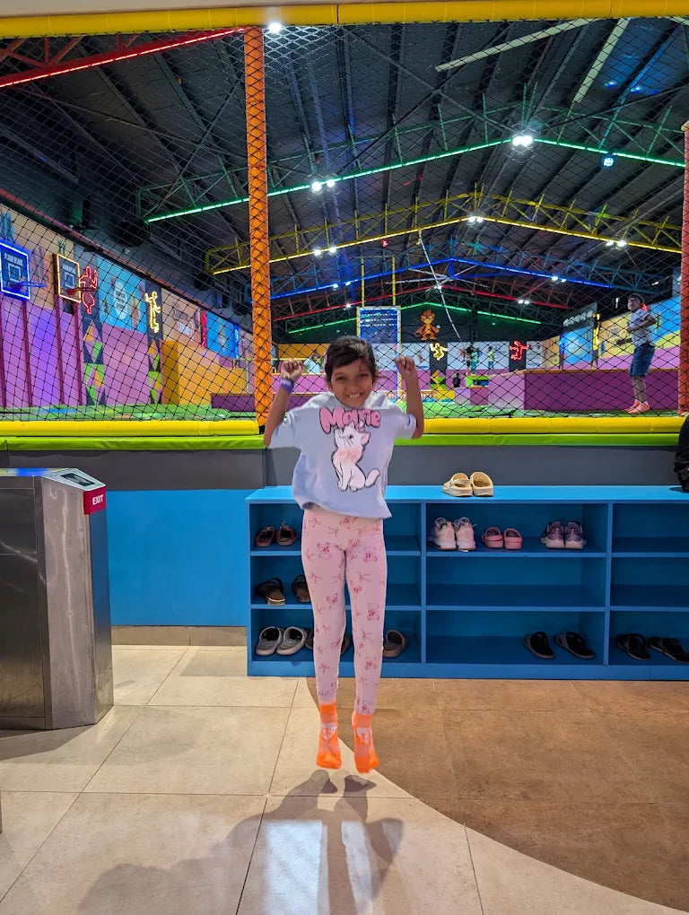 A young child jumps in the air at an indoor trampoline park, with colorful netting and lights in the background.