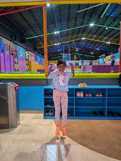 A young child jumps in the air at an indoor trampoline park, with colorful netting and lights in the background.