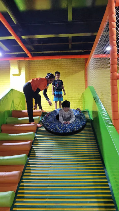 A woman helps children at an indoor playground slide. One child is in a tube at the bottom of the slide.