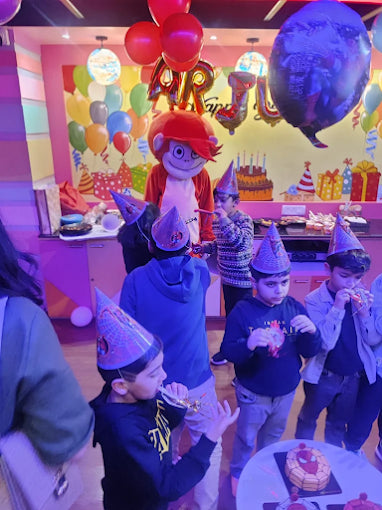 Children wearing party hats enjoying a colorful birthday celebration with balloons and a mascot in a decorated room