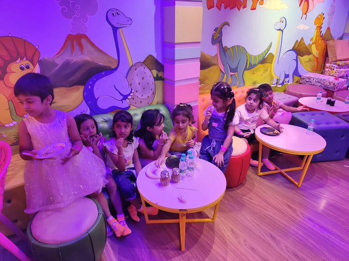 Children enjoying snacks and drinks in a colorful dinosaur-themed party room with playful wall murals and cozy seating