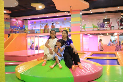 Three smiling girls sitting together on a colorful round platform in a bright indoor trampoline park play area