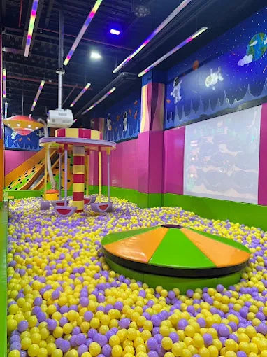 Indoor space-themed ball pit with yellow and purple balls and colorful climbing play equipment in a kids trampoline park