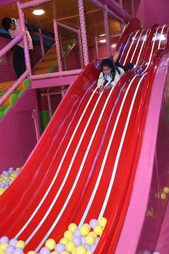 Child enjoying a bright red triple slide descending into a yellow and purple ball pit at indoor trampoline park