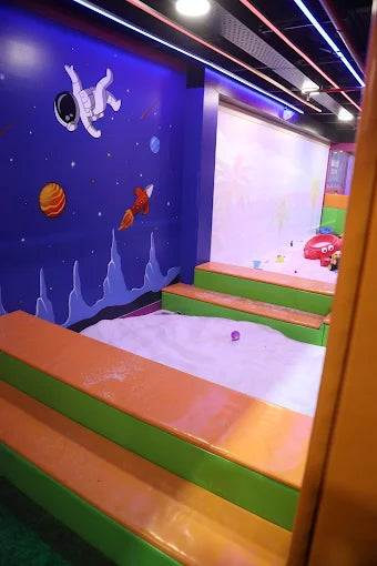 Kids indoor play zone with space-themed wall mural and soft seating surrounding a sand play area at a trampoline park