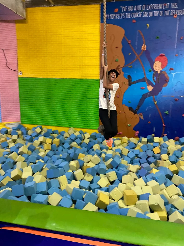 A young person laughs while climbing a rope over a foam pit at an indoor adventure park.