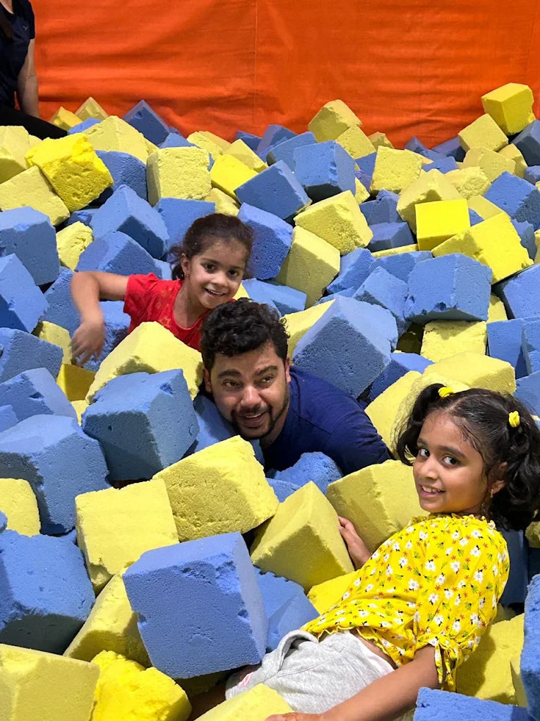 A father and two young girls play in a foam pit filled with blue and yellow cubes.