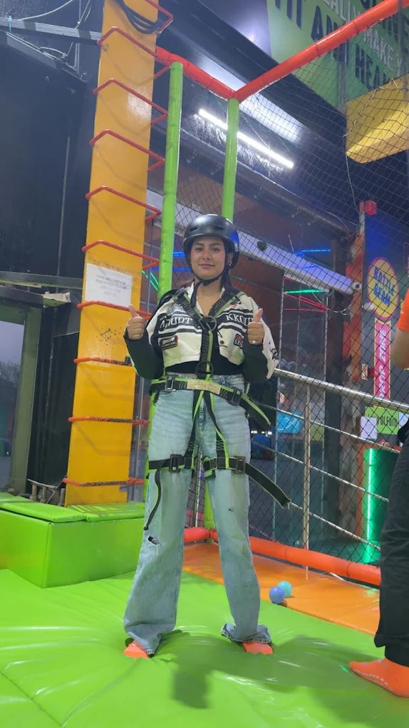 A young woman in a helmet and harness gives a thumbs-up on a colorful indoor adventure course.