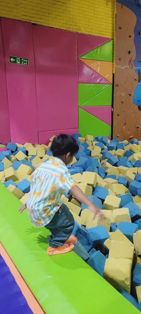A young child jumps into a foam pit at an indoor play area. The child wears a striped shirt and is surrounded by blue and yellow foam blocks.