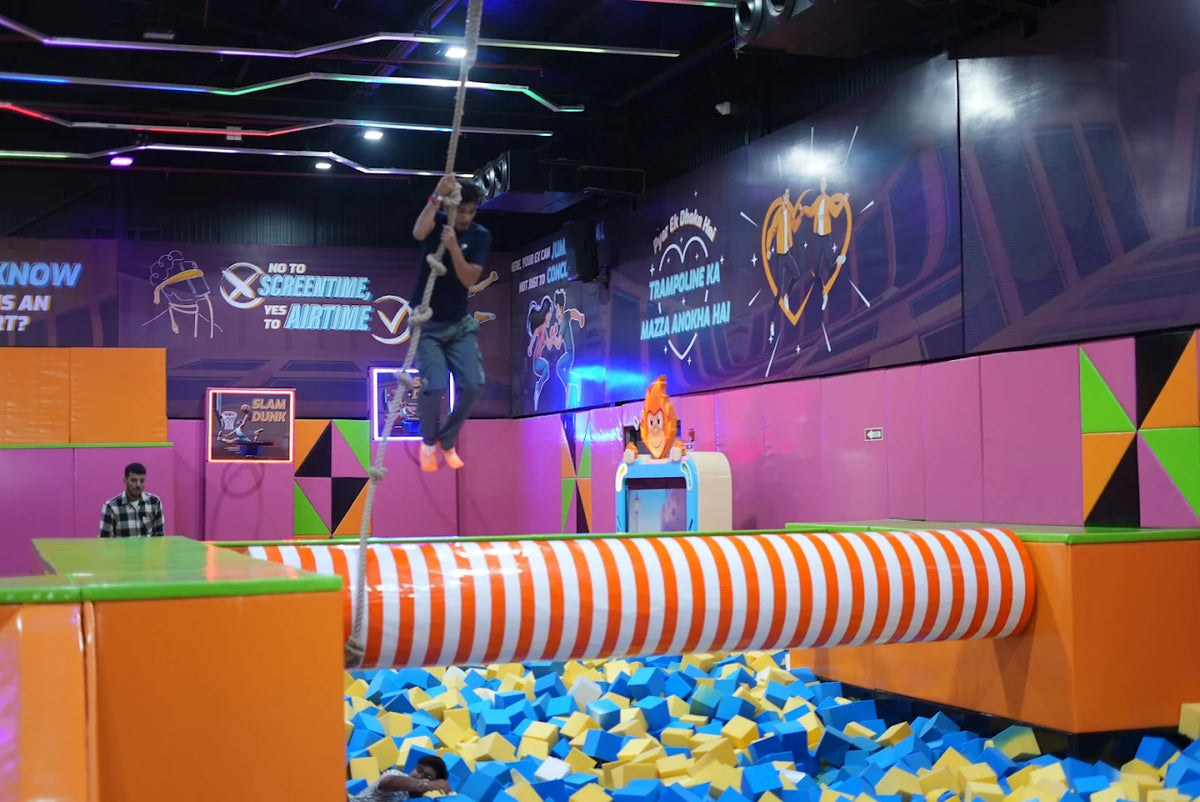 A young person swings on a rope over a foam pit at a trampoline park.
