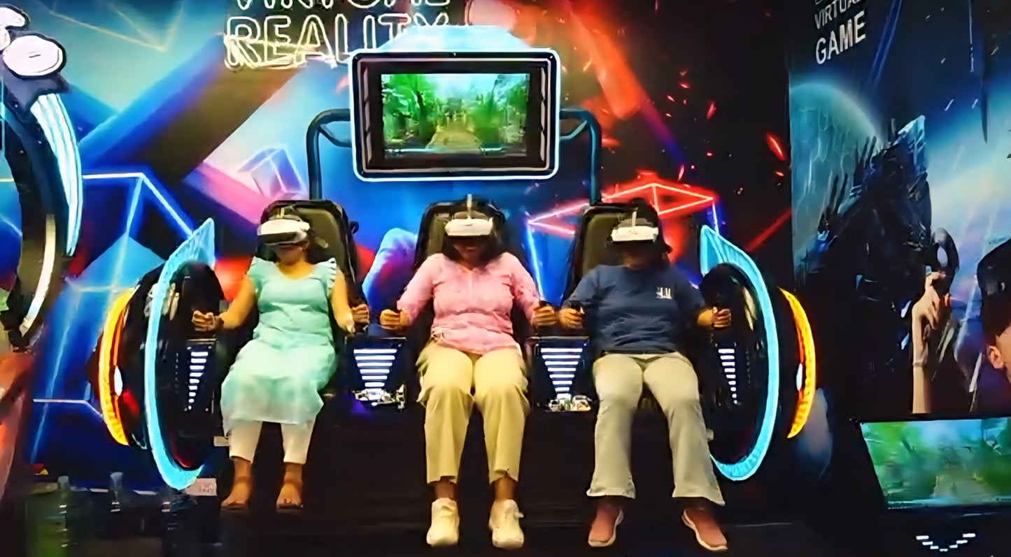 Three adults wearing VR headsets sit in a virtual reality game simulator. The setting is a brightly lit arcade with neon lights.