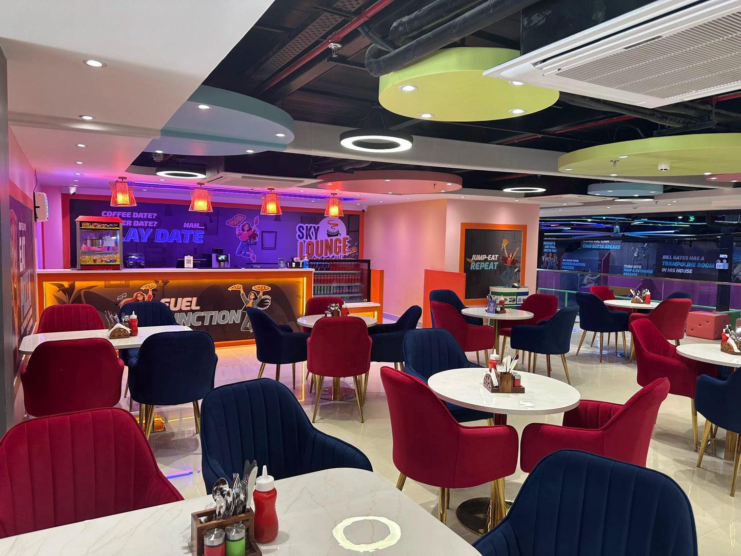 A modern cafe with red and blue velvet chairs and tables, featuring a popcorn machine and colorful decor.