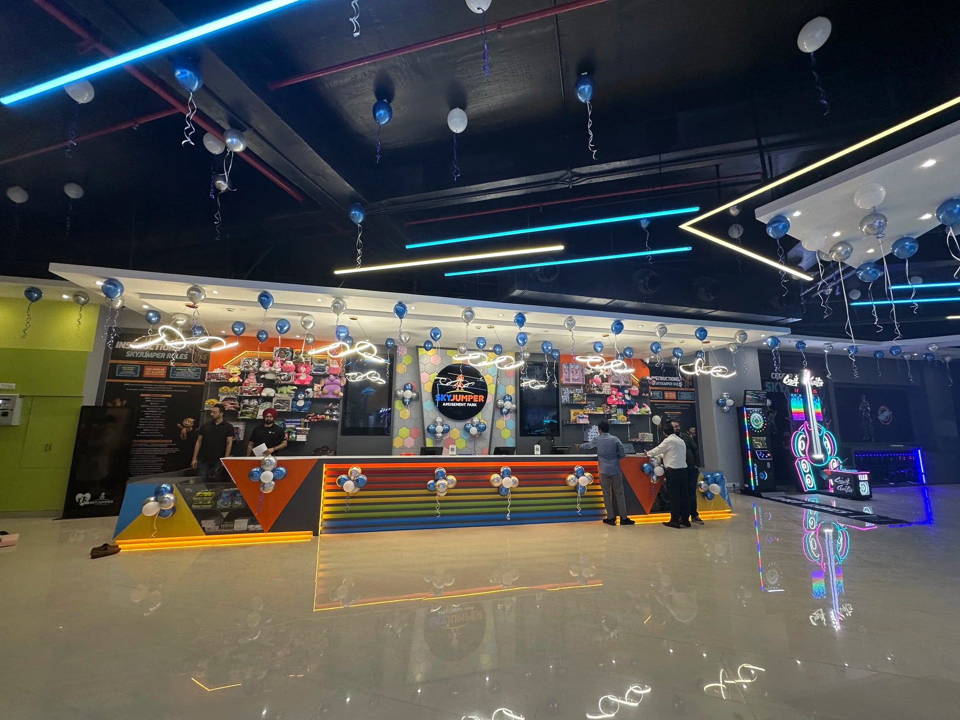 People at an amusement park with blue and silver balloons and neon lights.