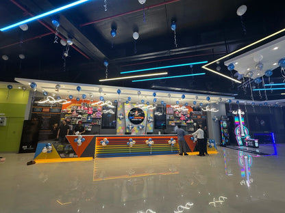 People at an amusement park with blue and silver balloons and neon lights.