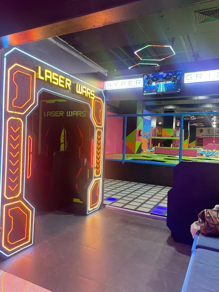 Entrance to a laser tag arena with neon lights and a trampoline park in the background.
