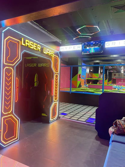 Entrance to a laser tag arena with neon lights and a trampoline park in the background.
