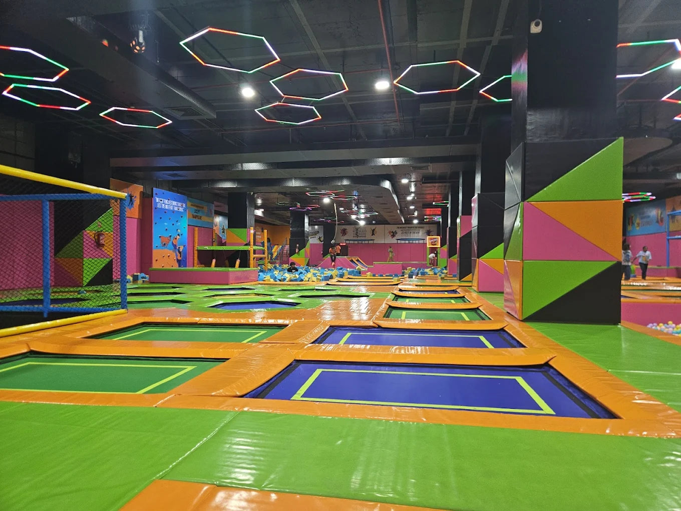 An indoor trampoline park with many trampolines and colorful padding, with a few children jumping.