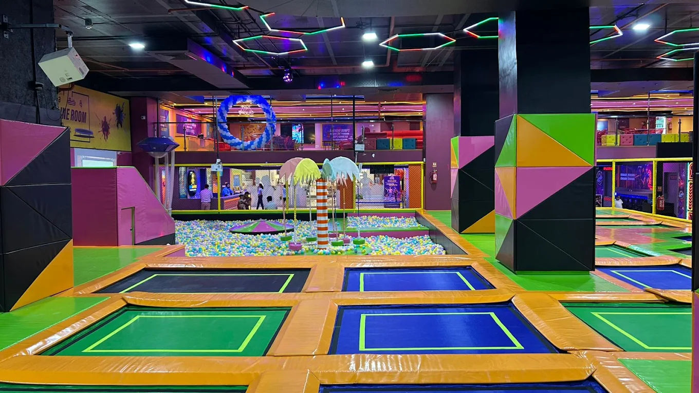 An indoor trampoline park with colorful trampolines, a ball pit, and geometric decorations, filled with children and young adults.