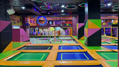 An indoor trampoline park with colorful trampolines, a ball pit, and geometric decorations, filled with children and young adults.
