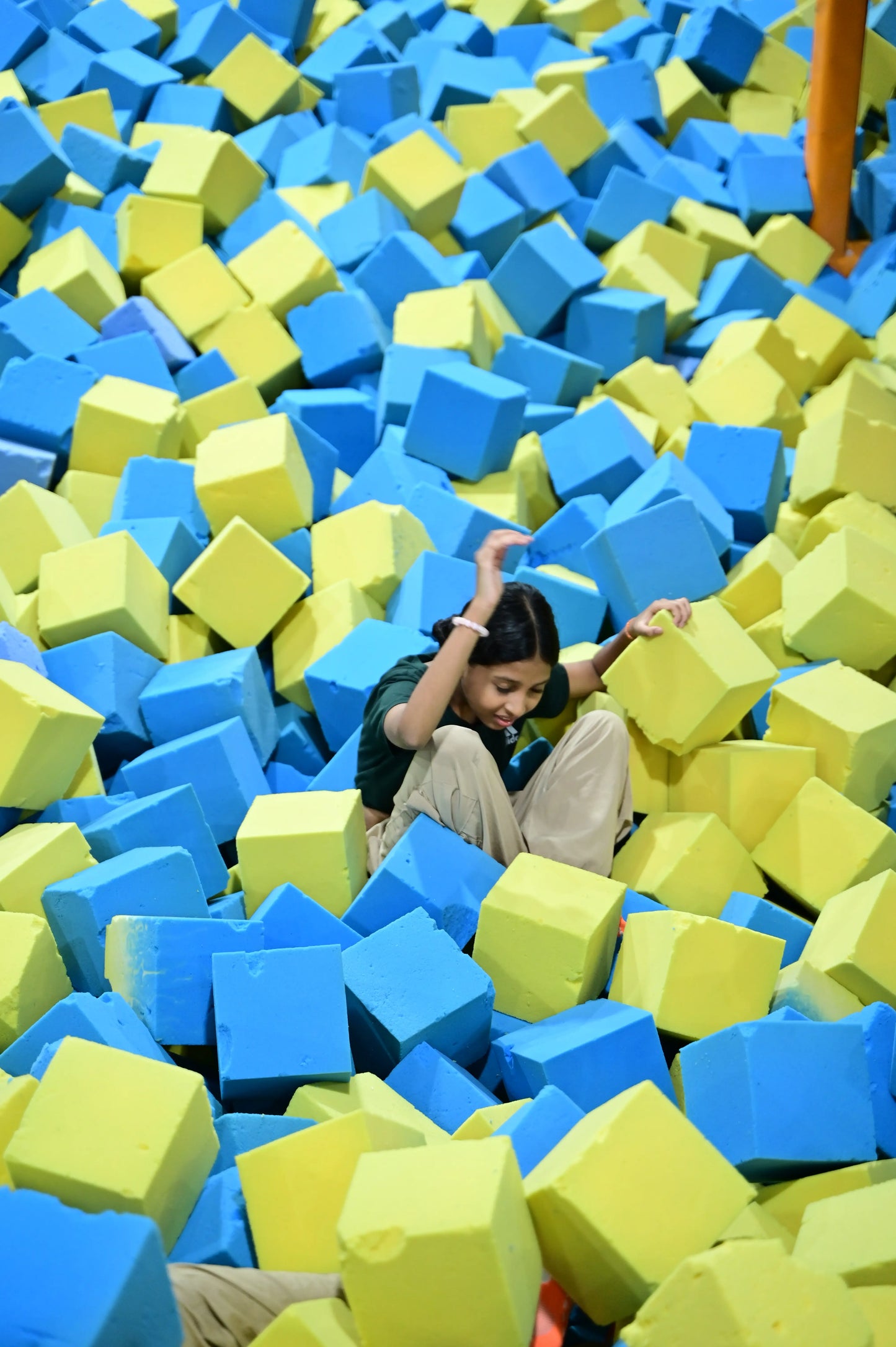 A young person sits in a pit of blue and yellow foam cubes, looking down.