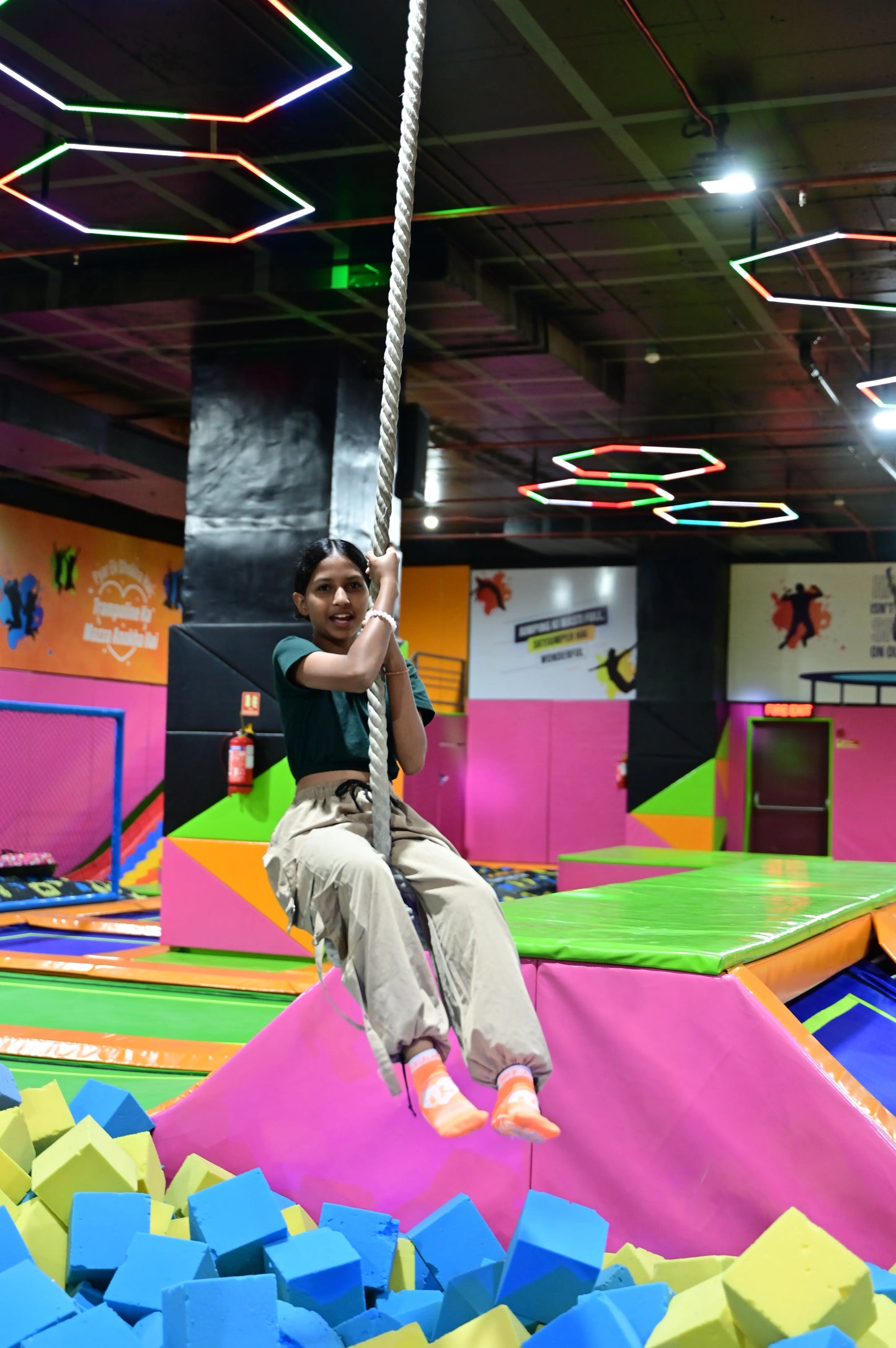 A young person swings on a rope over a foam pit in a colorful indoor trampoline park.