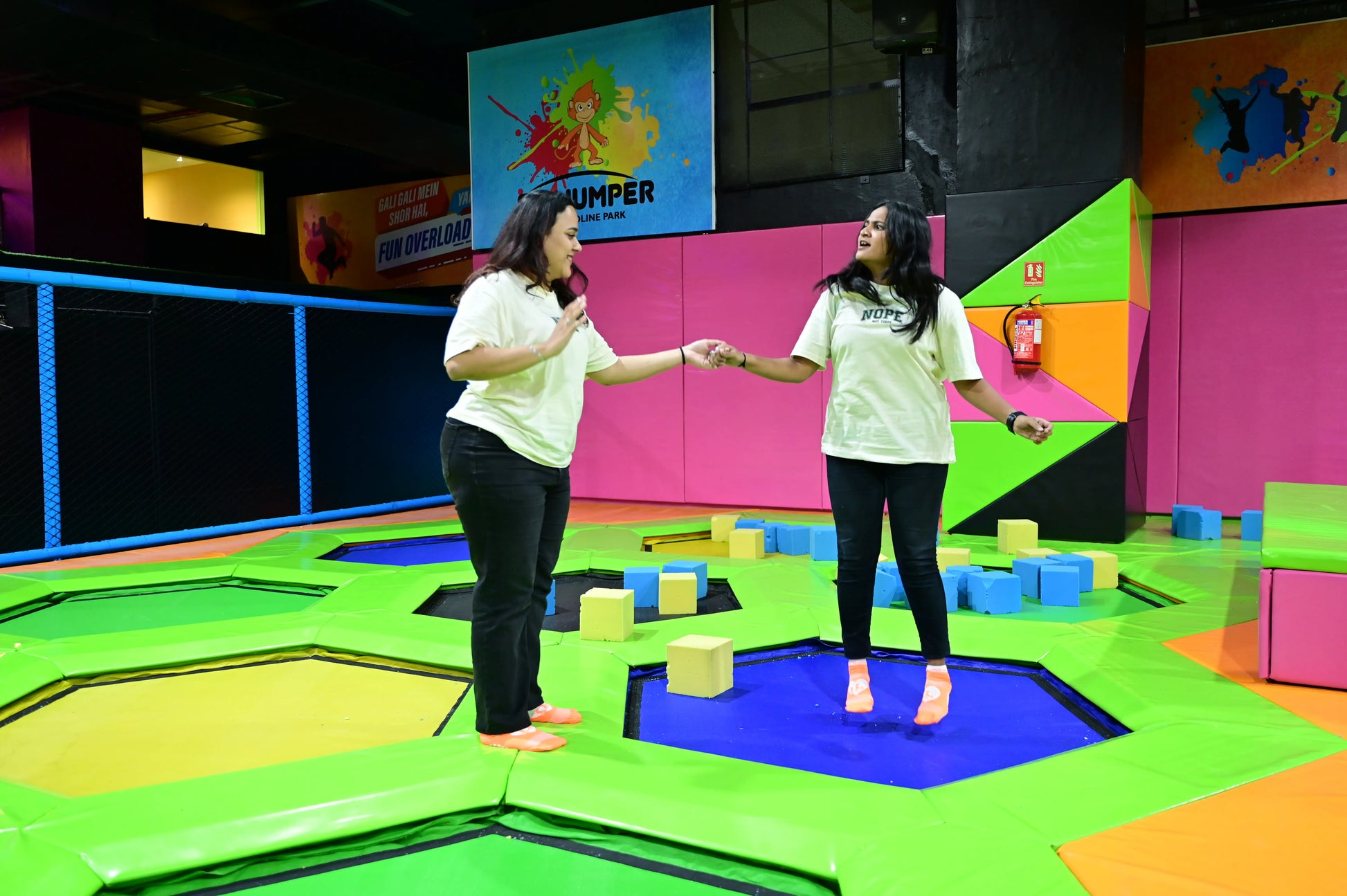Two young women hold hands while jumping on trampolines in a colorful indoor park.