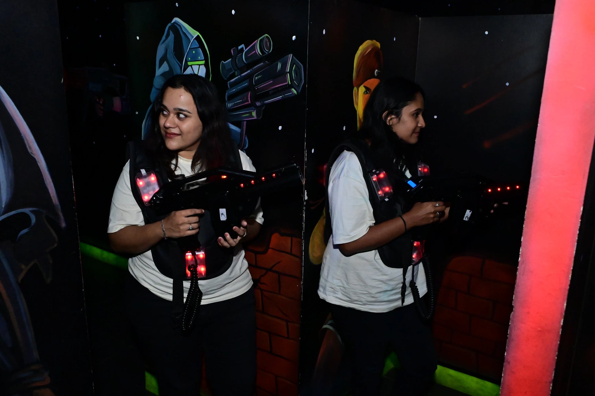 Two young women play laser tag in a dimly lit arena with futuristic murals. They wear vests and hold laser guns.