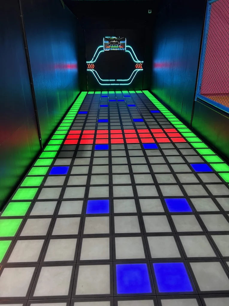 An arcade game floor with glowing blue, red, and green tiles, leading to a neon-lit screen.