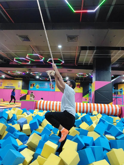 A young man climbs a rope over a foam pit at a trampoline park.
