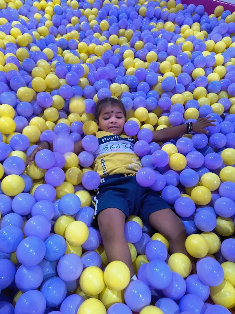 A young child lies in a ball pit filled with yellow and purple balls.