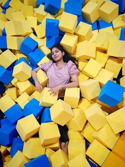 A young woman lies in a ball pit filled with yellow and blue foam cubes.