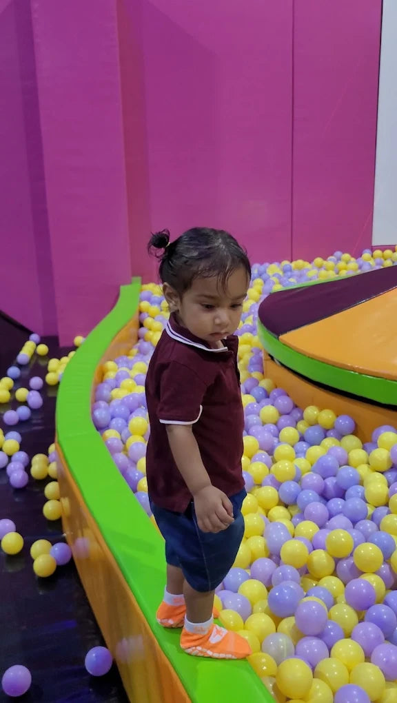 A young child stands on the edge of a ball pit filled with purple and yellow balls.