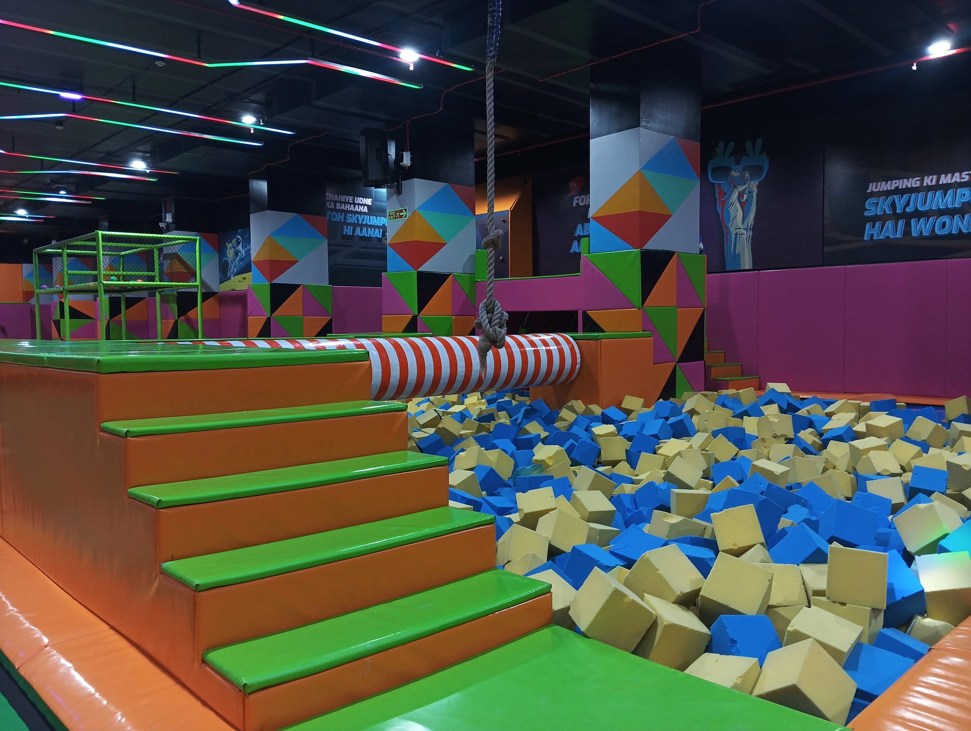 A foam pit filled with blue and yellow cubes, with colorful padded stairs leading to a trampoline area.