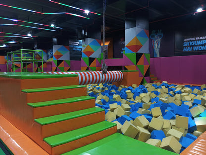 A foam pit filled with blue and yellow cubes, with colorful padded stairs leading to a trampoline area.