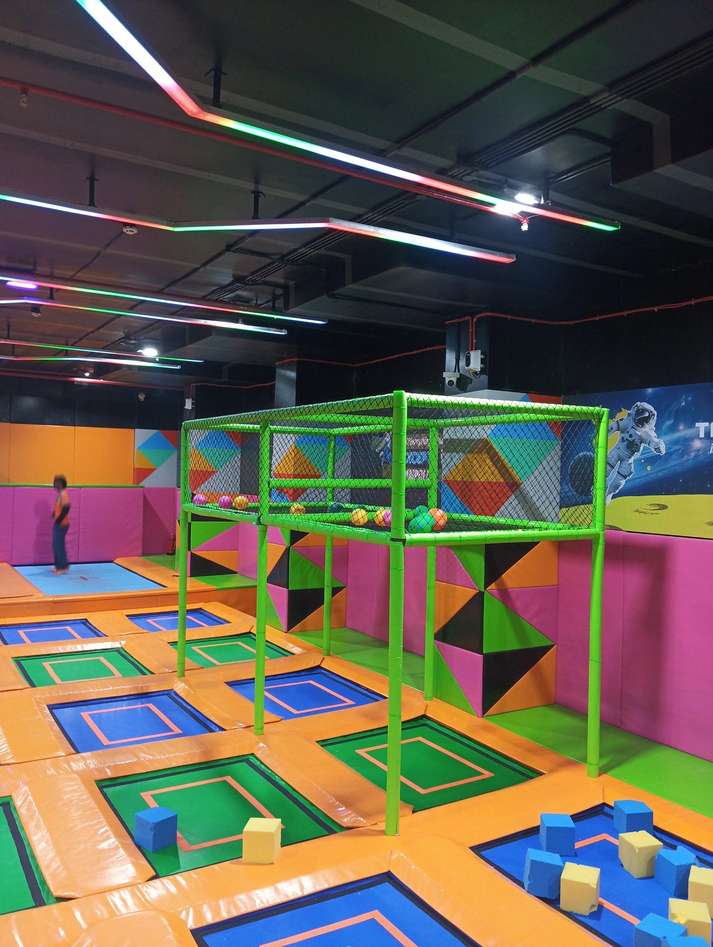 A colorful indoor trampoline park with a person jumping and foam cubes scattered on trampolines.