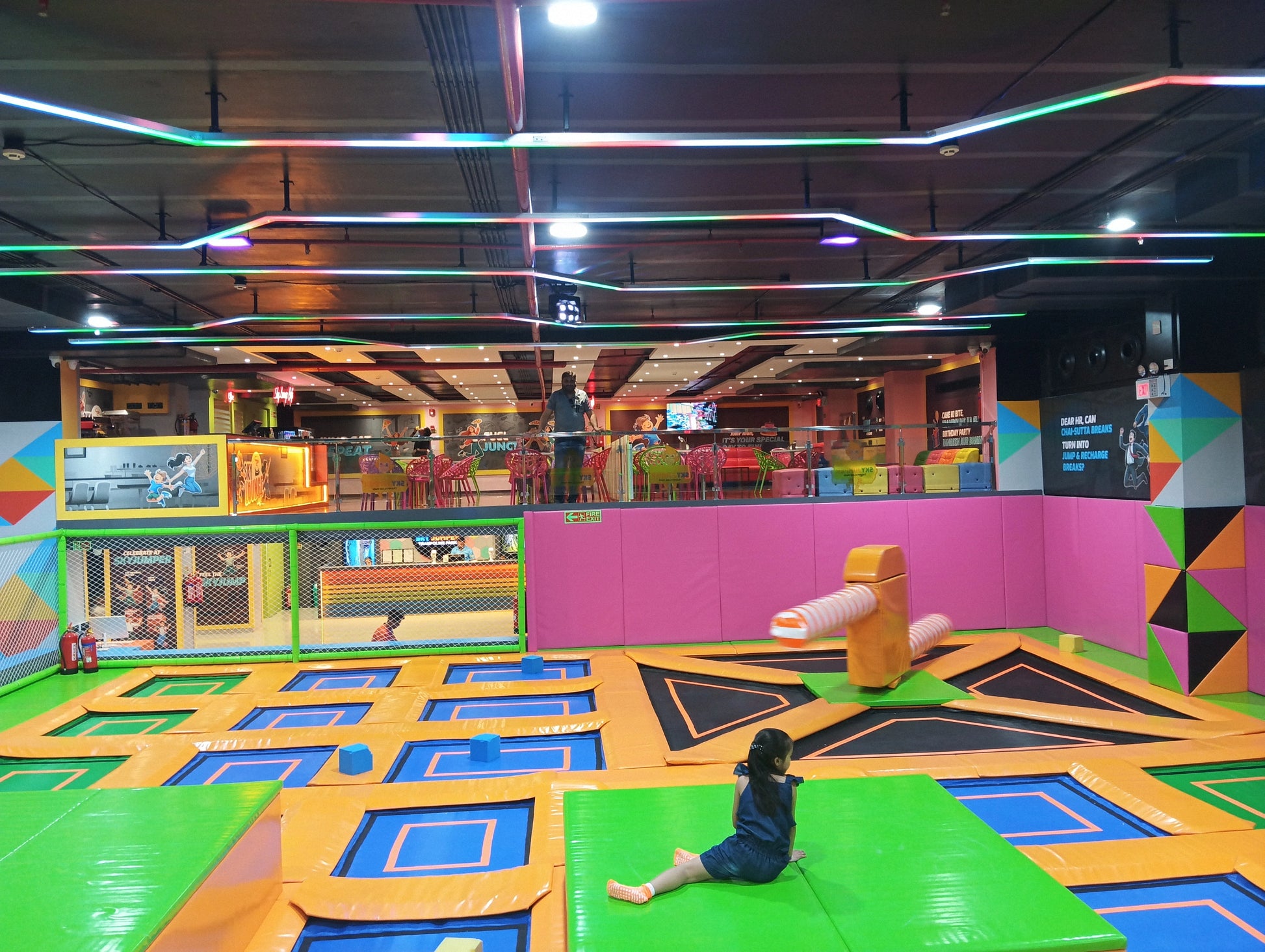 A young girl sits on a trampoline at an indoor trampoline park with colorful lights overhead.