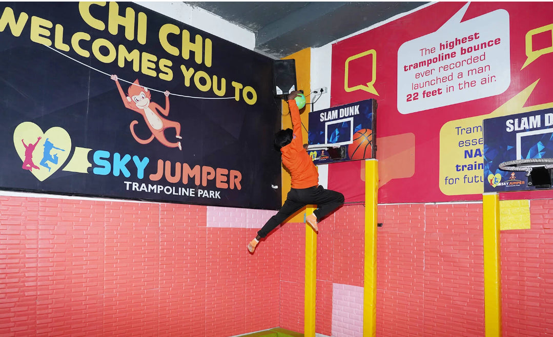 A young person in an orange shirt jumps to dunk a basketball at a trampoline park.