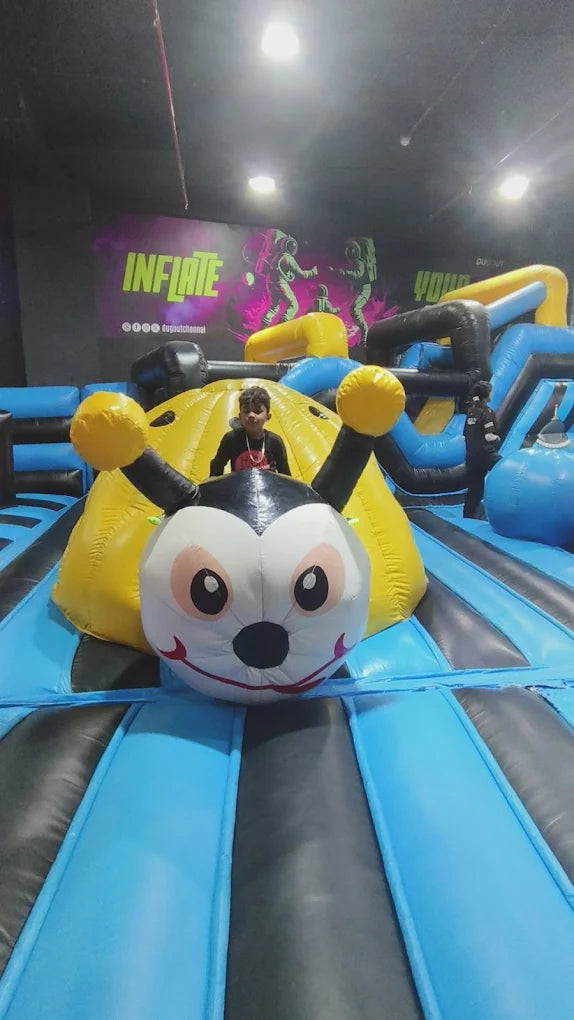 A young boy sits on a large inflatable bee at an indoor playground. The playground features bright blue and yellow inflatable structures.