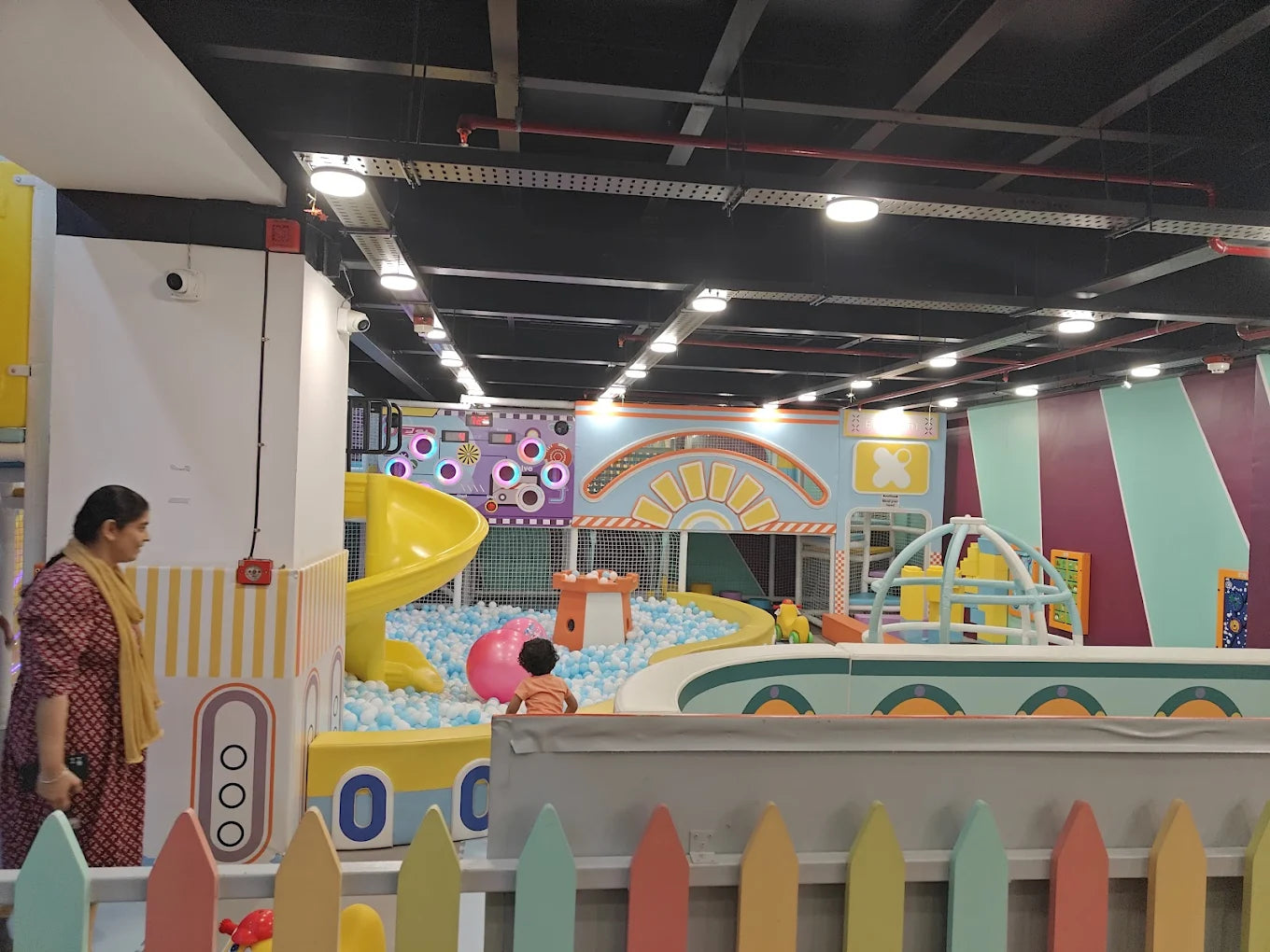 A child plays in a ball pit at an indoor playground, with a woman standing nearby.
