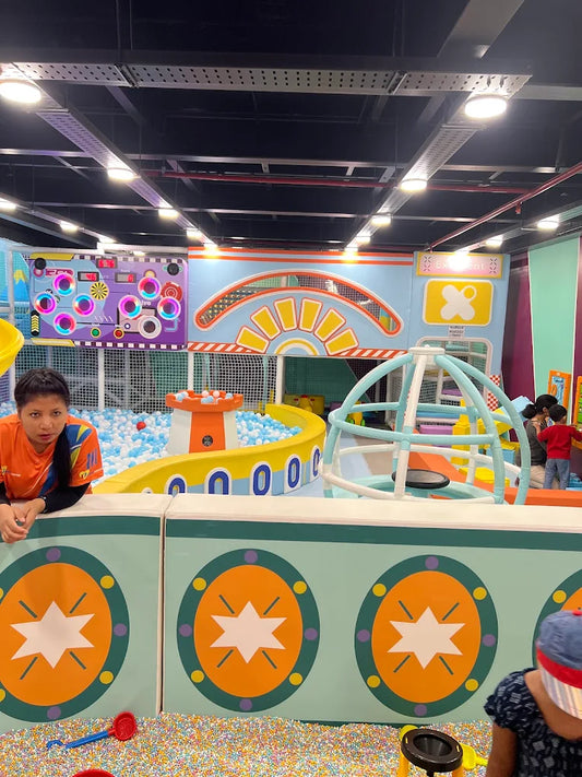 A woman watches children play in a colorful indoor playground with a ball pit and climbing structure.