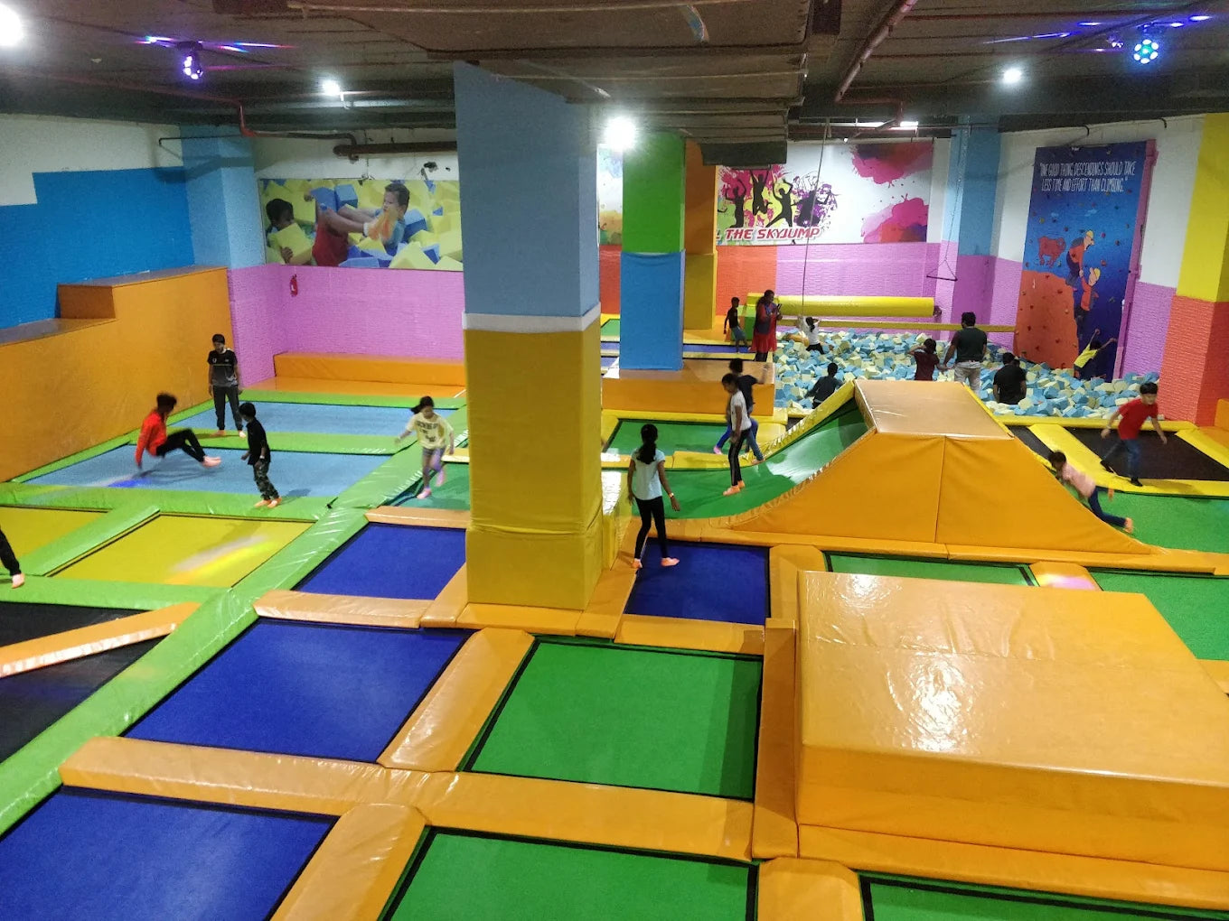 Children jump on trampolines in an indoor play area with foam pits and colorful walls.