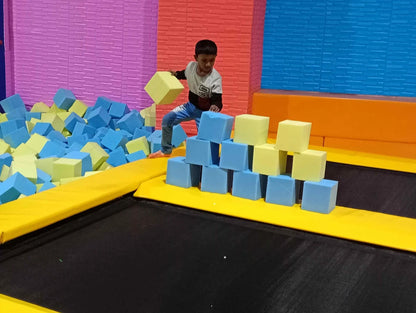 A young boy plays in a foam pit filled with colorful blocks, stacking them on a trampoline.