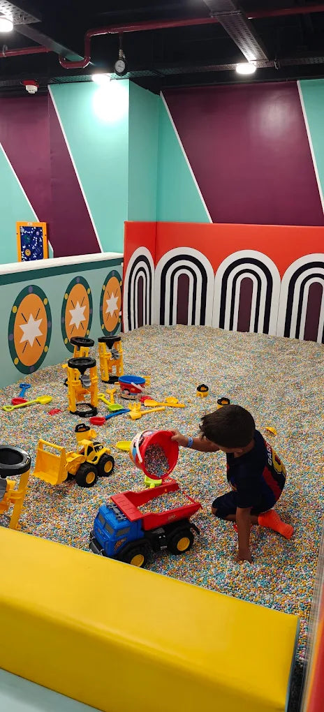 A young child plays with colorful toys in a ball pit. The child is pouring small balls from a red bucket into a toy truck.