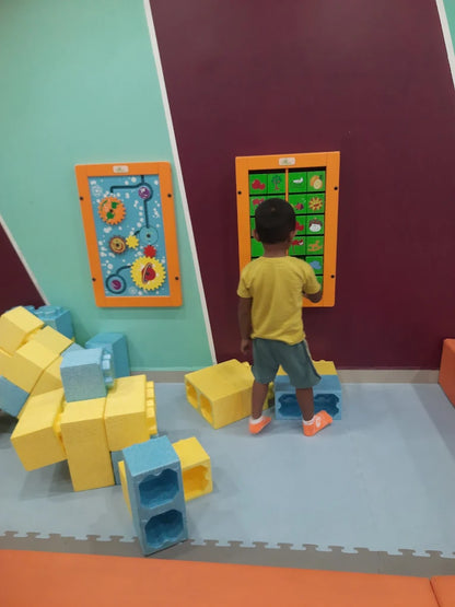 A young child plays with large foam blocks in a colorful room with interactive wall panels.