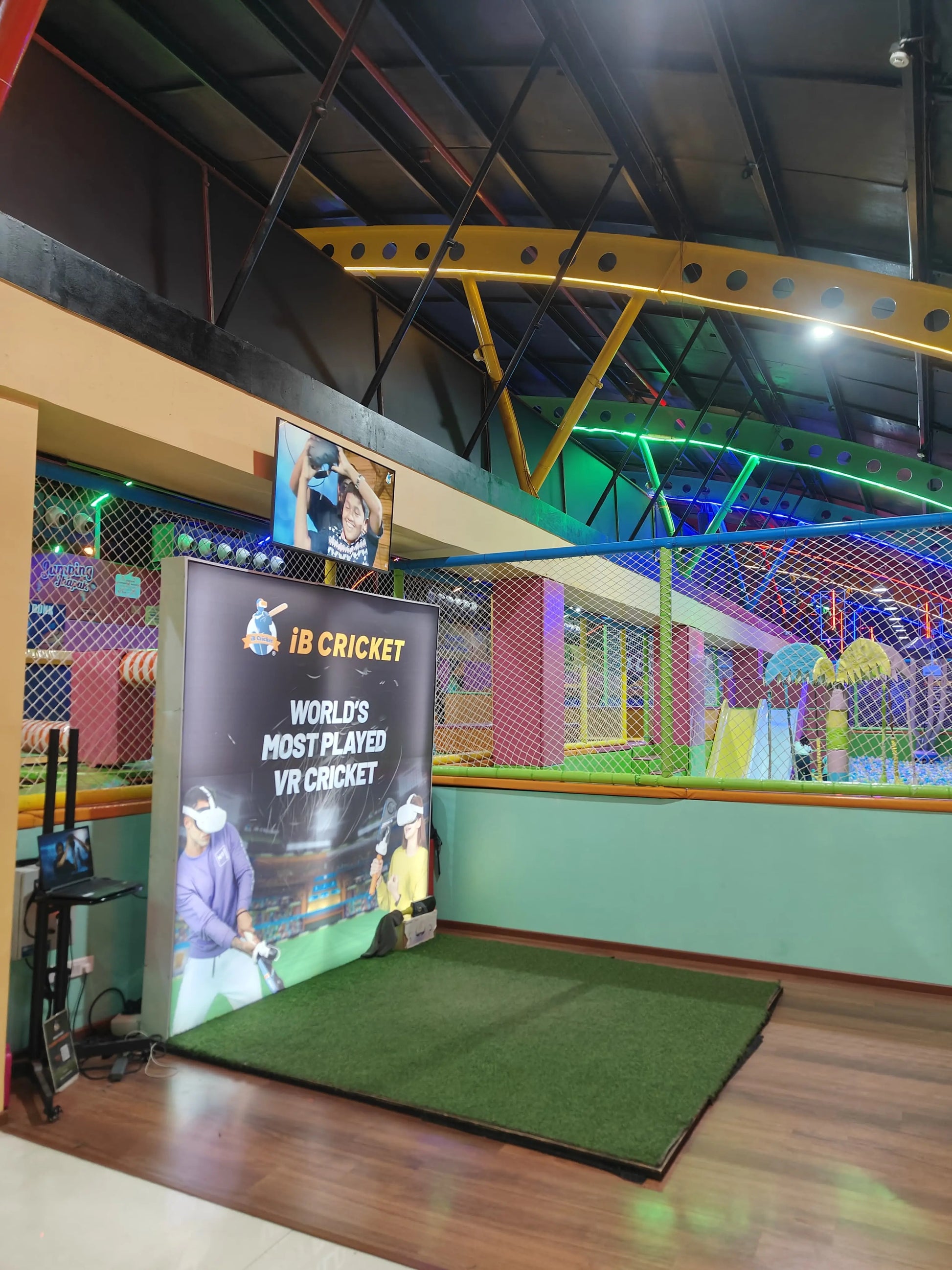 A VR cricket setup with a large banner and a young person playing. The background shows a colorful indoor playground.