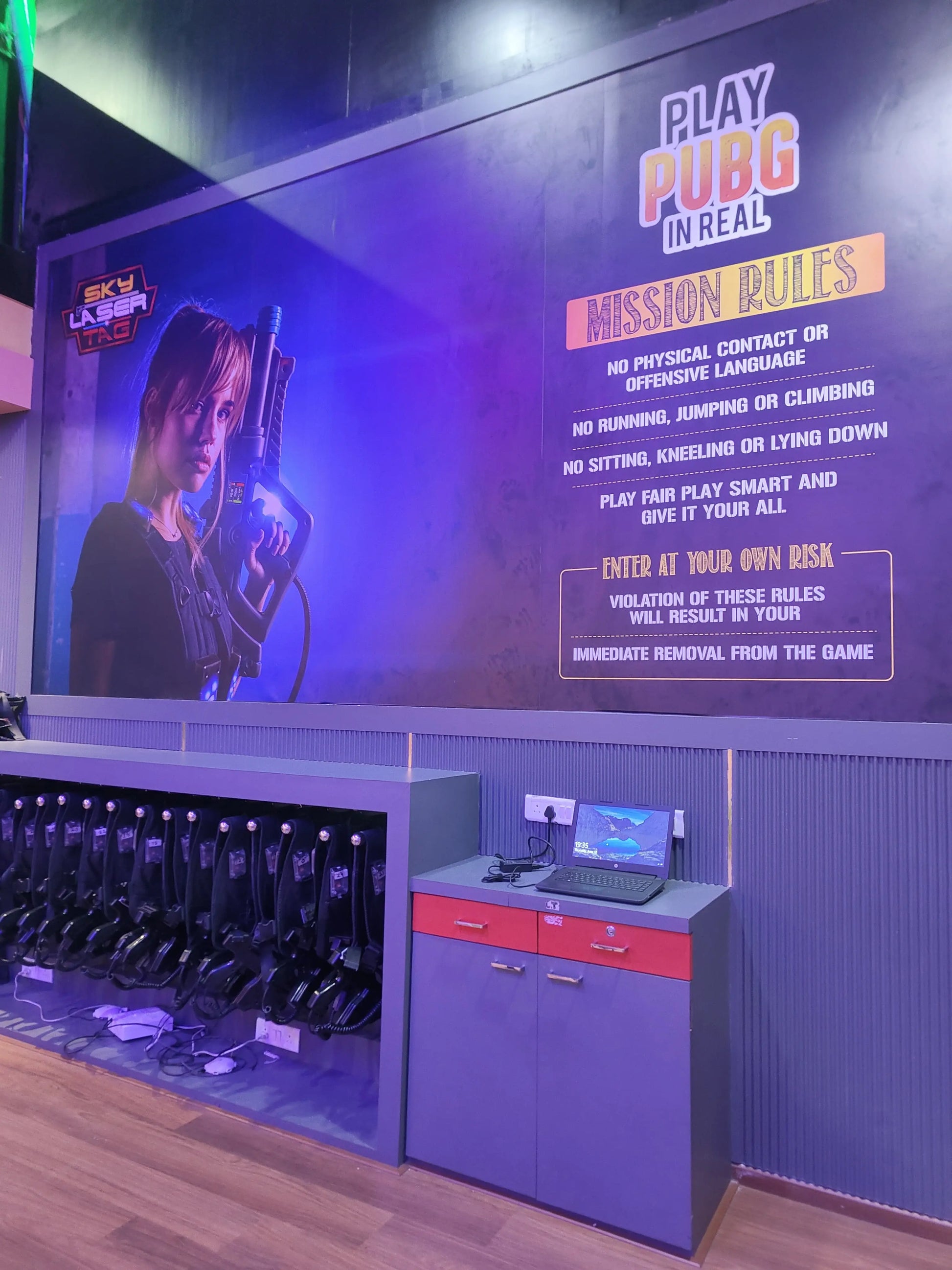 A laser tag arena with rules posted on a wall and vests hanging on display. A laptop sits on a counter next to the vests.