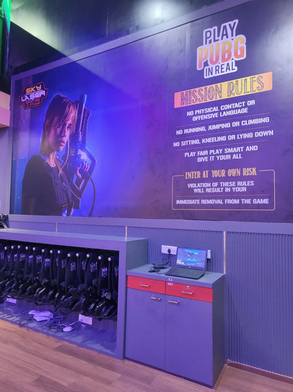 A laser tag arena with rules posted on a wall and vests hanging on display. A laptop sits on a counter next to the vests.