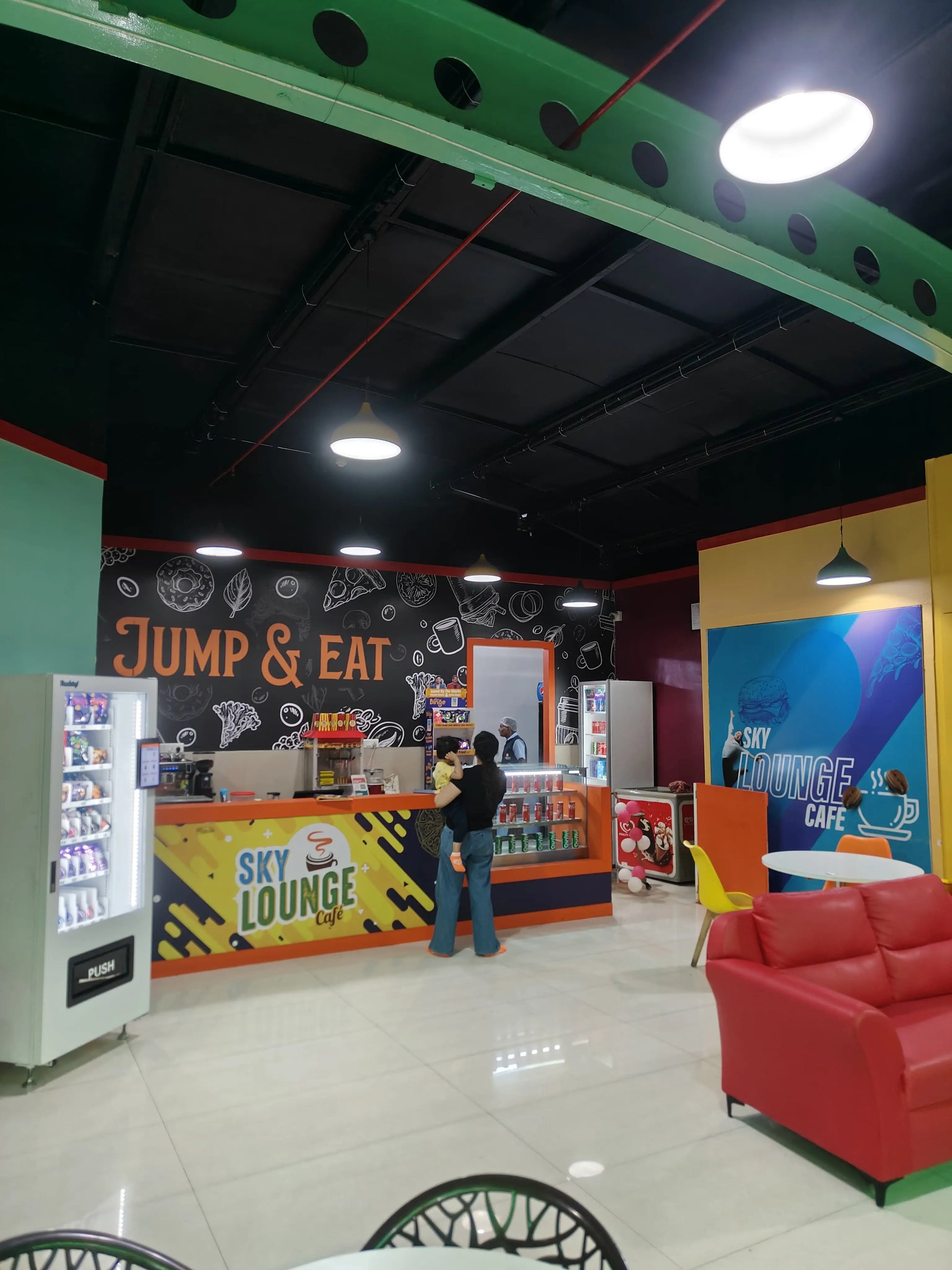 A woman holds a child at a cafe counter in a brightly lit indoor entertainment venue.